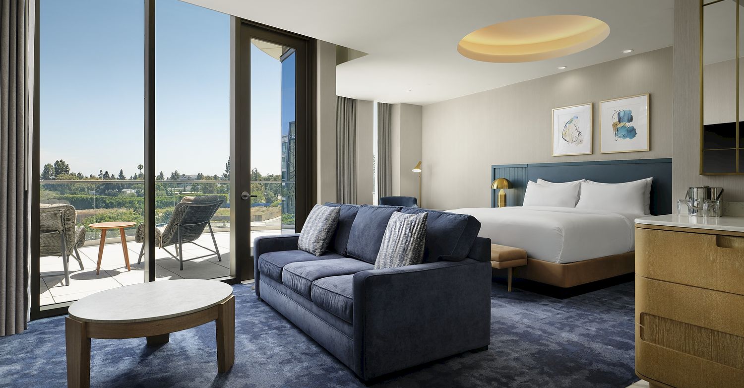 A modern hotel suite with a blue sofa, king bed, round table, large windows, and an outdoor terrace beyond them.