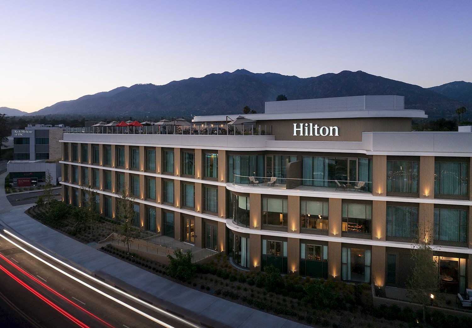 A Hilton hotel building with glowing windows, curving terrace, red lights on the road, and mountains in the background.