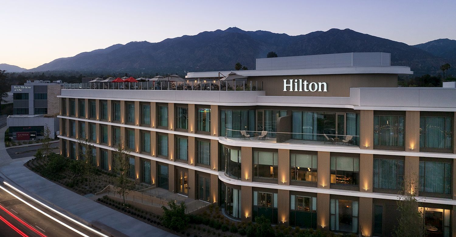 A Hilton hotel building with glowing windows, curving terrace, red lights on the road, and mountains in the background.