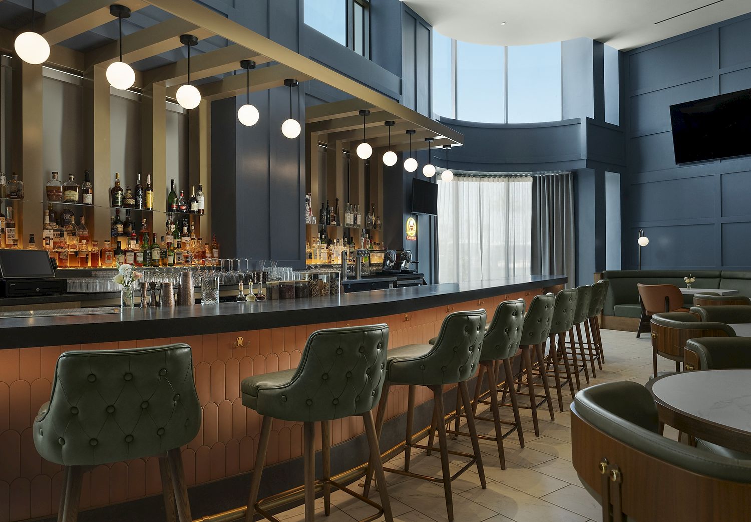 An elegant hotel bar with shelves of liquor, a long counter, hanging globe lights, and green tufted stools.