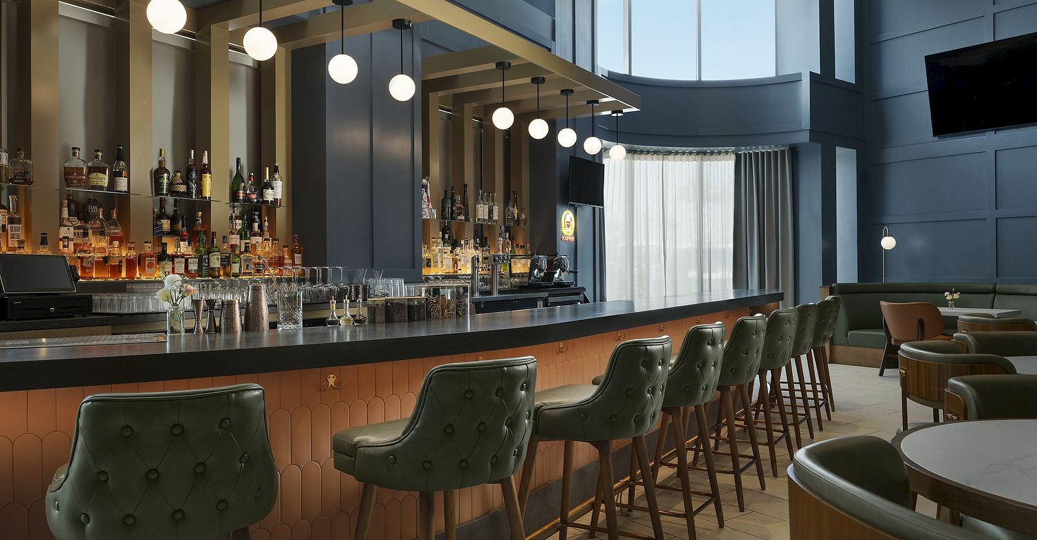 An elegant hotel bar with shelves of liquor, a long counter, hanging globe lights, and green tufted stools.