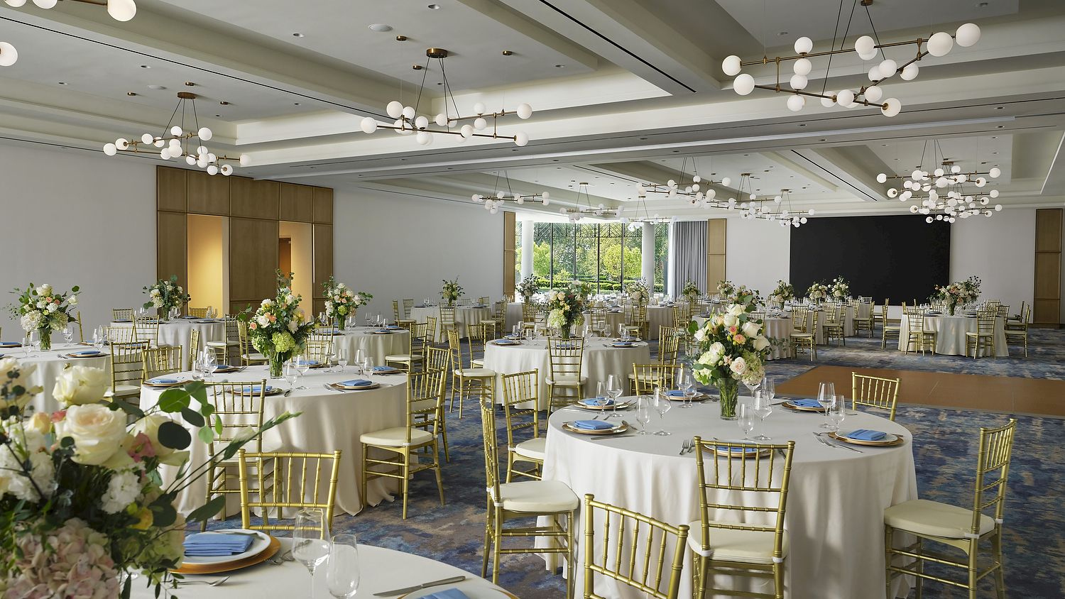 An elegant banquet hall set with white table linens, gold chairs, floral centerpieces, and sparkling globe chandeliers overhead.