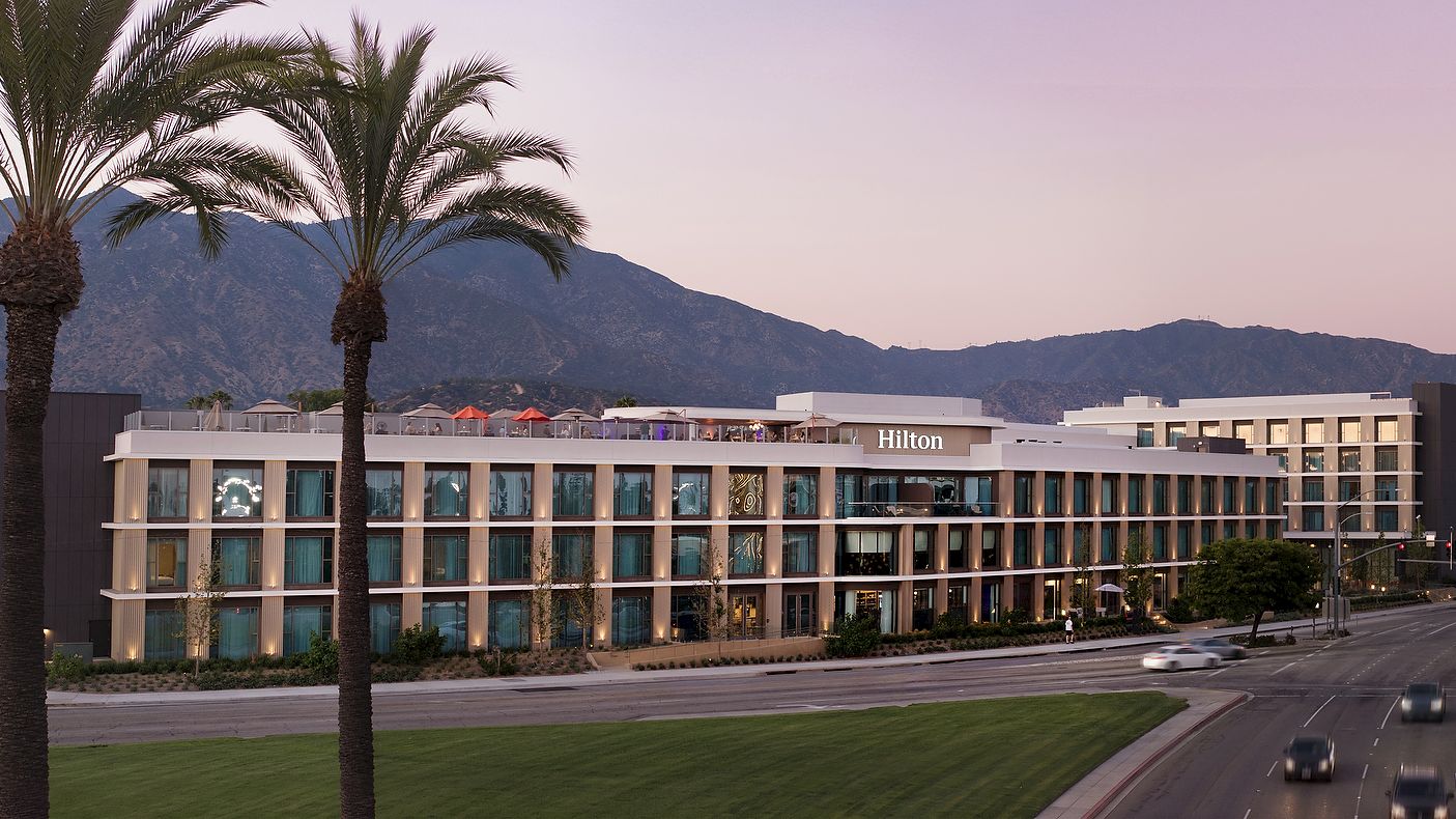 A Hilton hotel exterior at dusk with tall palm trees, a curved road, and mountains in the background.