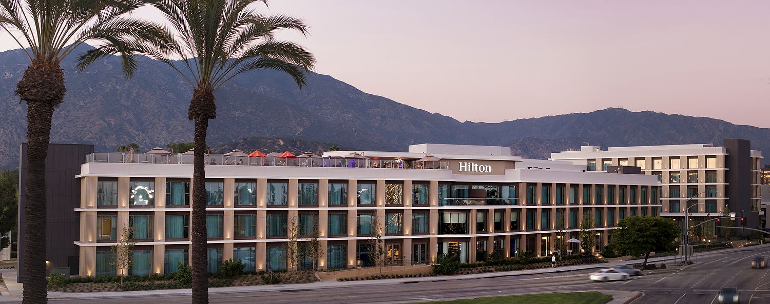 A Hilton hotel exterior at dusk with tall palm trees, a curved road, and mountains in the background.
