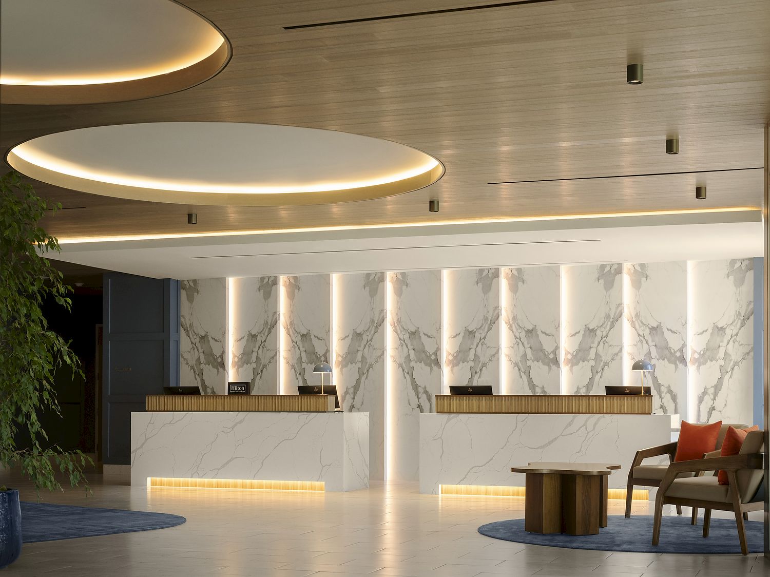 Modern hotel lobby with marble reception counters, warm circular ceiling lights, and a seating area with orange cushions.