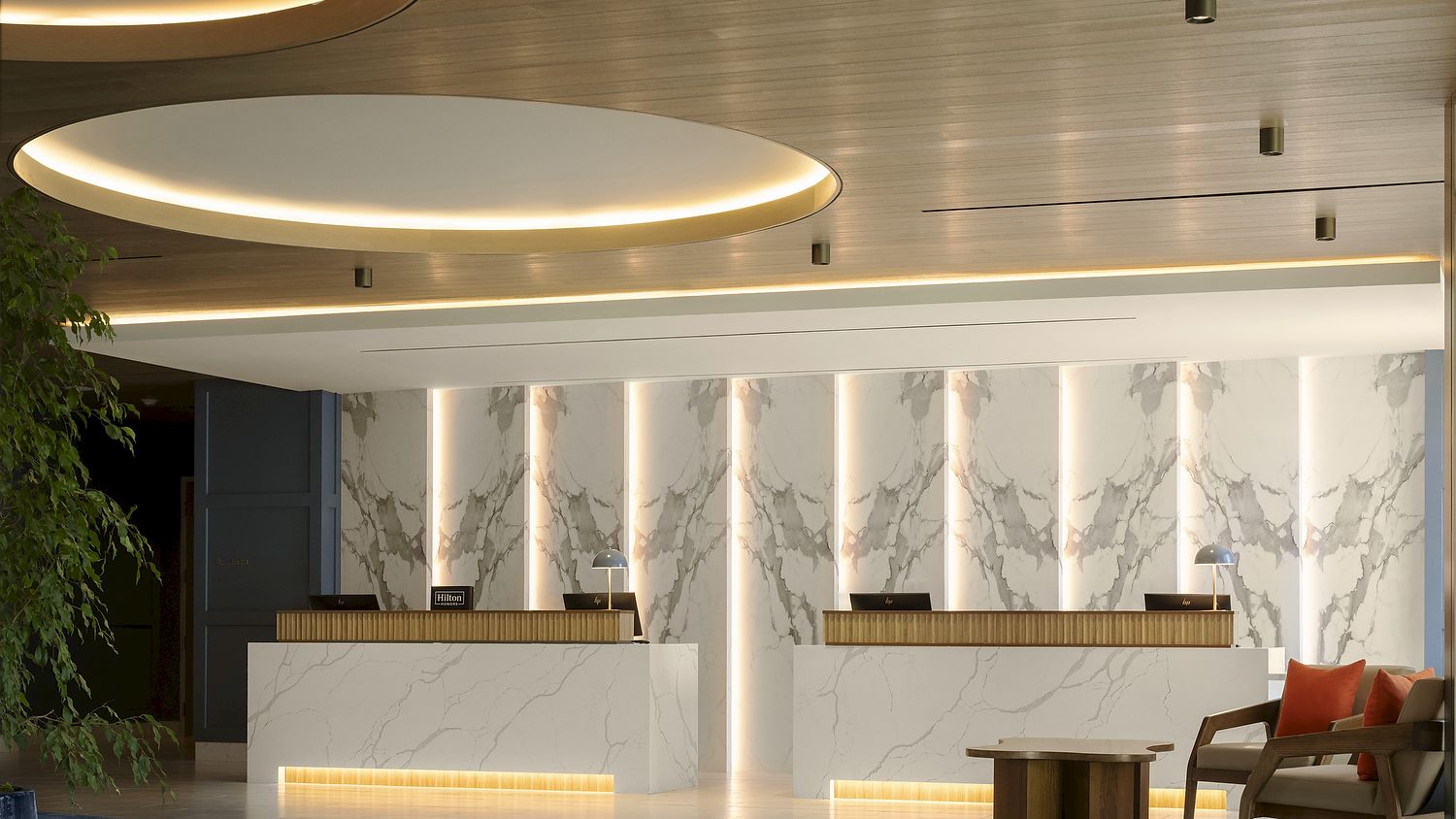 Modern hotel lobby with marble reception counters, warm circular ceiling lights, and a seating area with orange cushions.