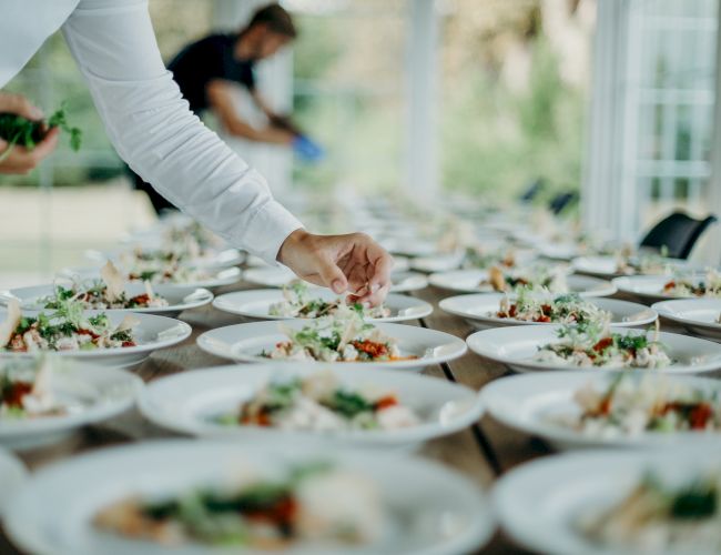 A server at an outdoor catered table places a garnished dish onto plates for guests to enjoy.