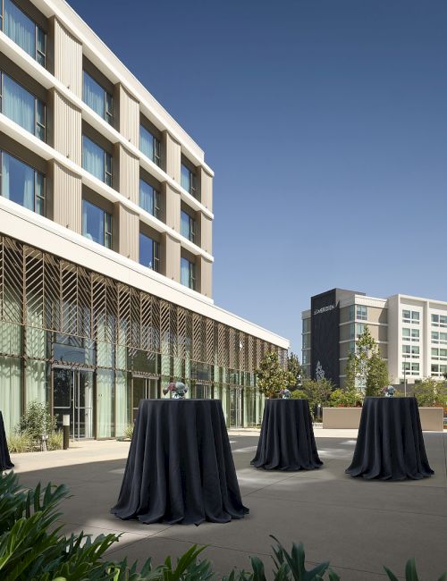 Modern hotel courtyard with tall buildings, outdoor tables in dark covers, potted flowers, and green plants under a blue sky.