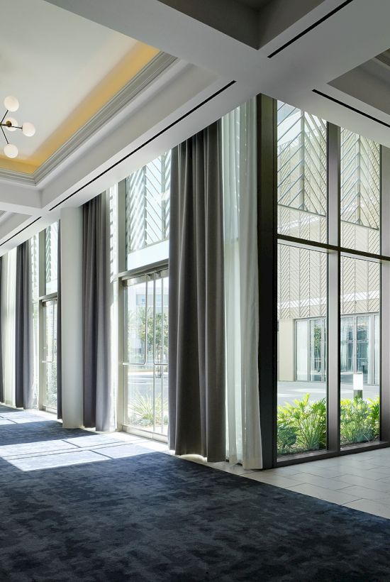 A sleek modern corridor with tall glass windows, dark curtains, hanging chandeliers, and indoor plants lining the hall.