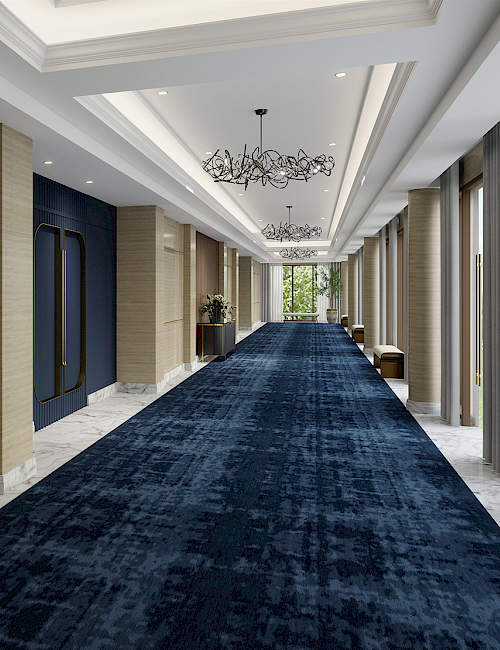 A modern hallway with a dark blue carpet, wooden panel walls, ceiling lights, and a chandelier, leading to a bright area.