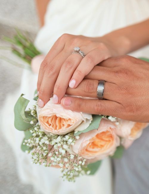 A couple's hands with wedding rings are placed over a bouquet of roses and baby's breath. The person on the right wears a watch.