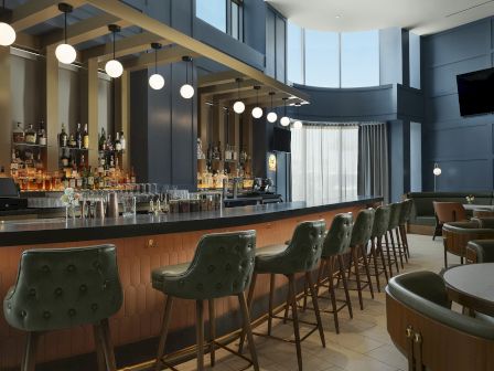 A modern hotel bar with bottles behind the counter, pendant lights, and green tufted stools and seating.