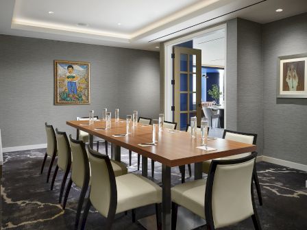 A modern conference room with a wooden table, six upholstered chairs, glass water carafes, and framed artwork on gray walls.