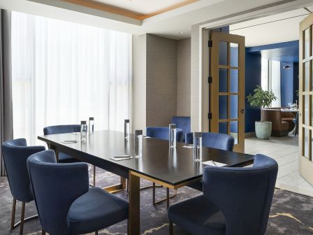 A modern conference room with a long black table, blue chairs, tall windows, and a doorway to a lounge area.