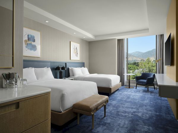 A modern hotel room with two beds, TV, desk, and a balcony view of mountains and greenery.