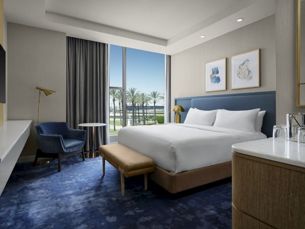A modern hotel bedroom with a king-size bed, blue carpet, curtains, framed art, and a view of palm trees.