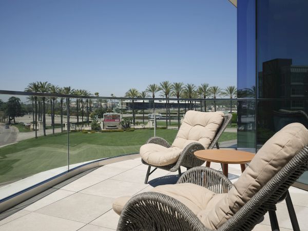 A balcony patio with wicker lounge chairs overlooks palm-lined grounds, a parking road, and distant resort buildings under a blue sky.