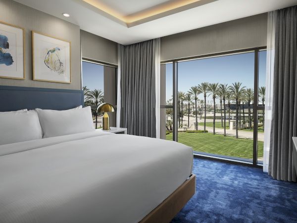 A modern hotel bedroom with a neatly made bed, floor-to-ceiling curtains, and windows overlooking palm-lined grounds.