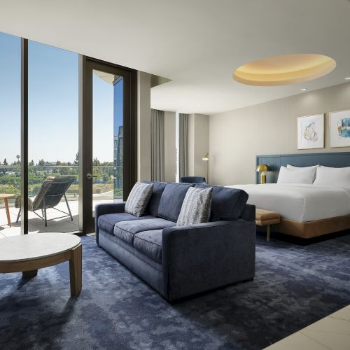 A modern hotel suite with a blue sofa, king bed, warm ceiling light, and open terrace view outside.