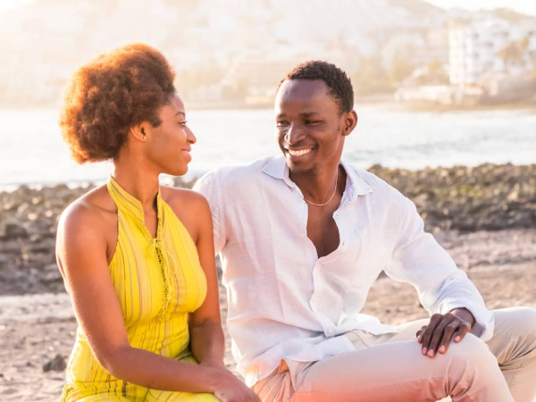 A couple is sitting on a beach, smiling at each other, with a body of water and a building in the background.