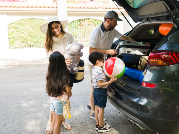 A family prepares for a trip, loading a car with beach gear, a teddy bear, and a beach ball while the kids watch.