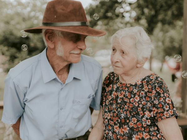 An elderly couple is standing outside. The man wears a brown hat and blue shirt; the woman is in a floral dress.