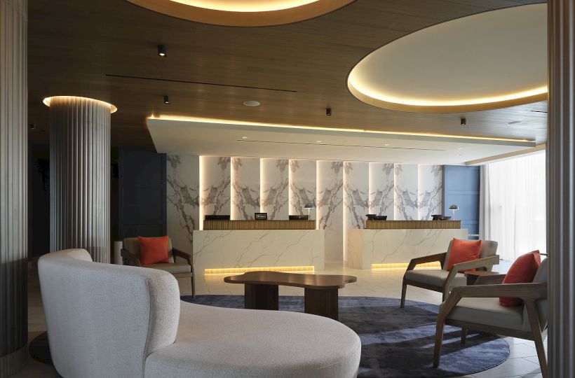 A modern hotel lobby with a marble front desk, warm recessed lighting, and comfortable lounge seating under curved ceiling lights.
