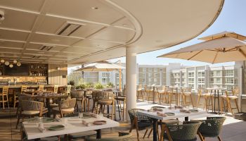 Sunlit patio restaurant with wooden chairs, table settings, and beige umbrellas beside a bar under a curved ceiling.