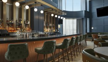 A modern hotel bar with shelves of liquor, hanging globe lights, and green tufted stools and lounge seating.