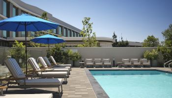 Sunny hotel courtyard with a blue swimming pool, loungers, and blue umbrellas under a clear sky.