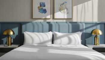 A modern bedroom with two framed abstract artworks above a blue paneled headboard, a white bed with pillows, and gold bedside lamps on either side.