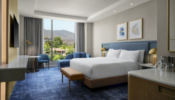 A modern hotel bedroom with a king bed, blue chairs, desk, framed art, and curtains opening to lush mountains.