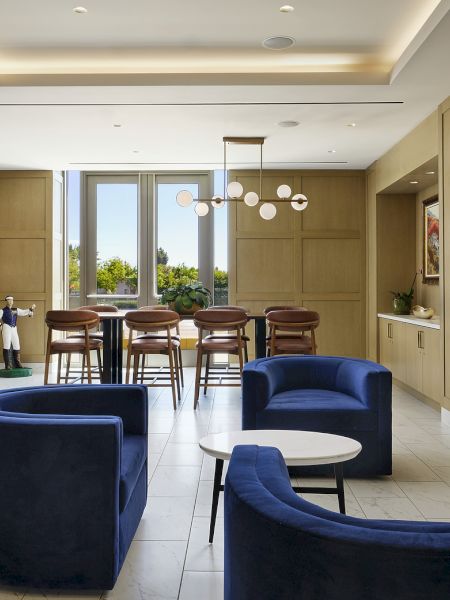 A modern hotel lounge with blue armchairs and sofas, wooden panel walls, bar stools, pendant lights, and large windows.