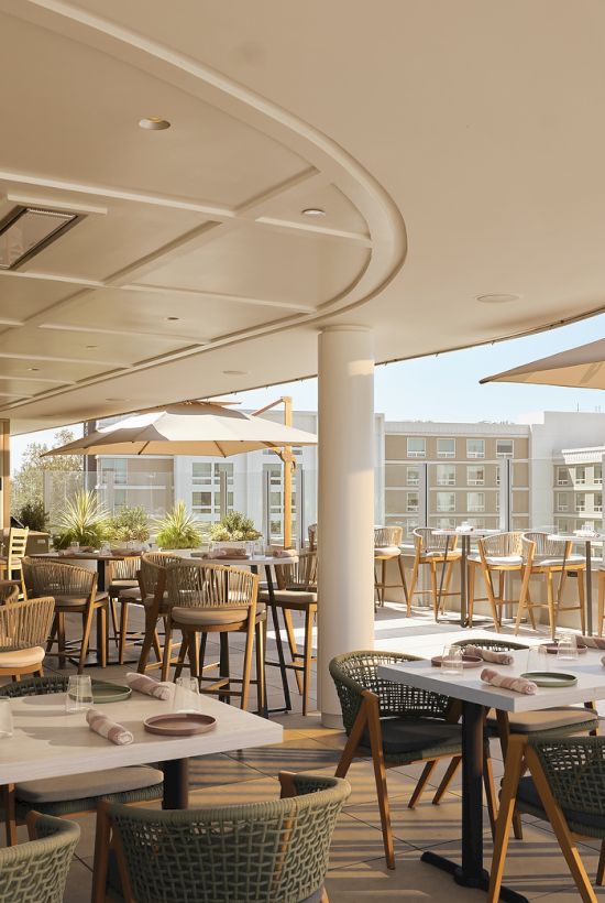 A hotel rooftop patio with tables, wicker chairs, umbrellas, and a bar, overlooking bright apartment buildings.