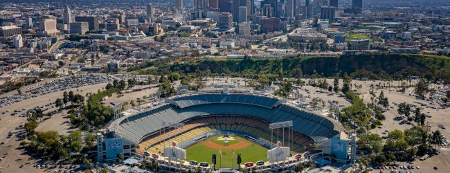 Aerial view of a large baseball stadium with a city skyline in the background, surrounded by parking lots and greenery.
