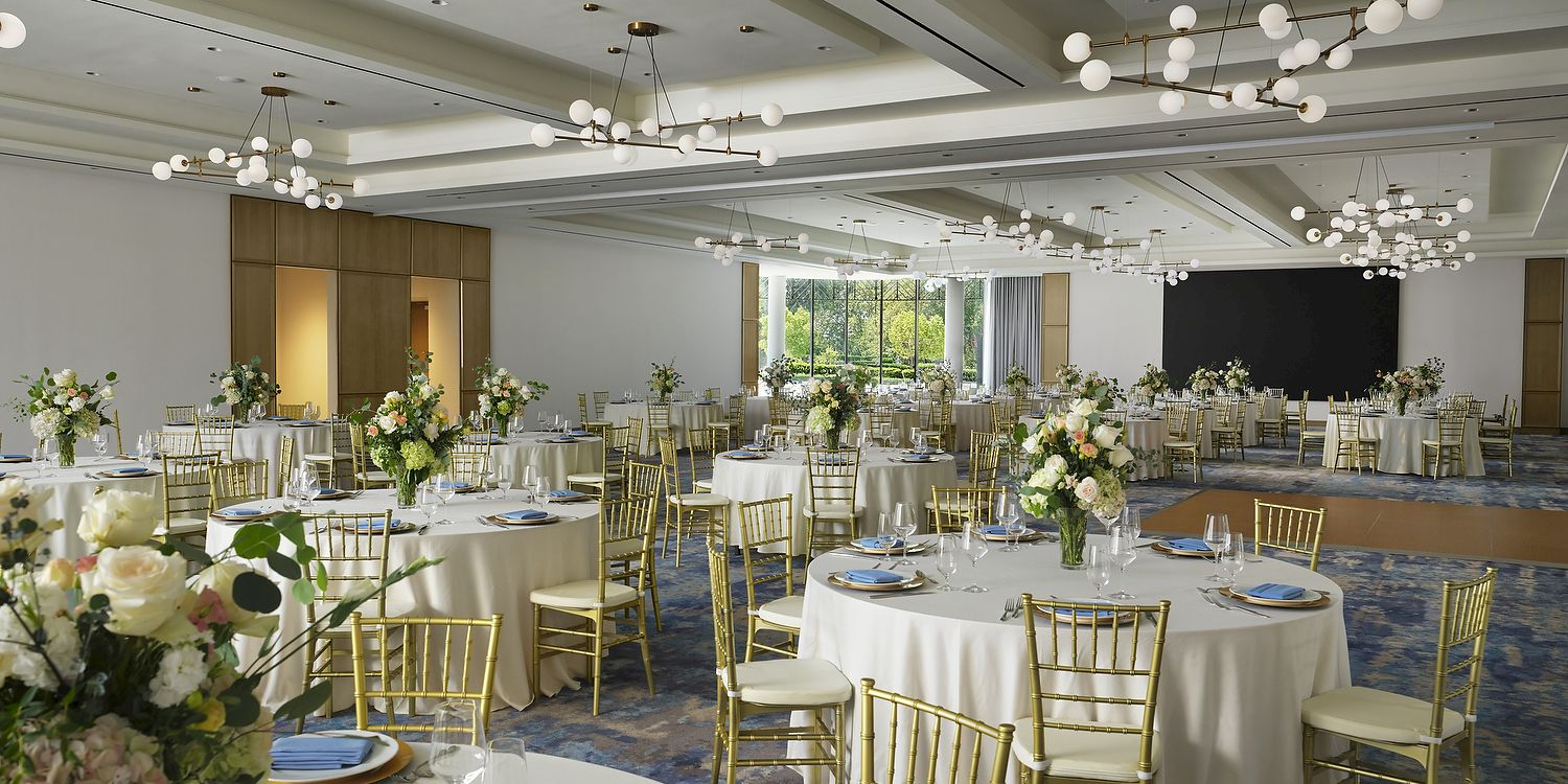 A bright event hall set for a wedding or banquet, with round tables, gold chairs, floral centerpieces, and globe chandeliers.