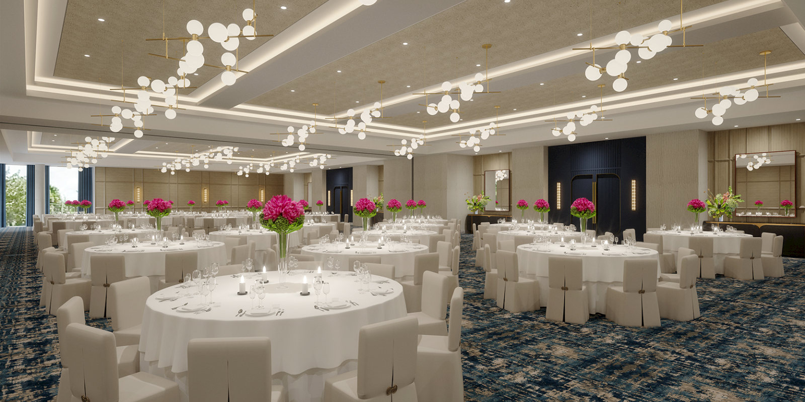 A banquet hall with round tables, white tablecloths, pink floral centerpieces, and elegant lighting on a patterned carpet.
