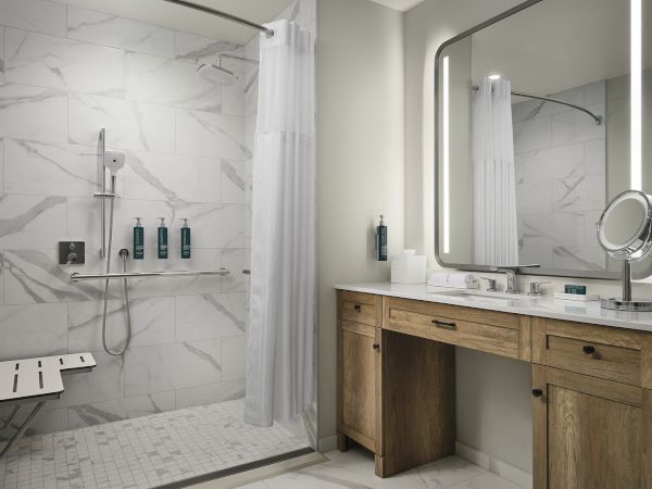 Modern bathroom with marble tiles, a glass-enclosed shower, white curtains, and a wooden vanity with an illuminated mirror.