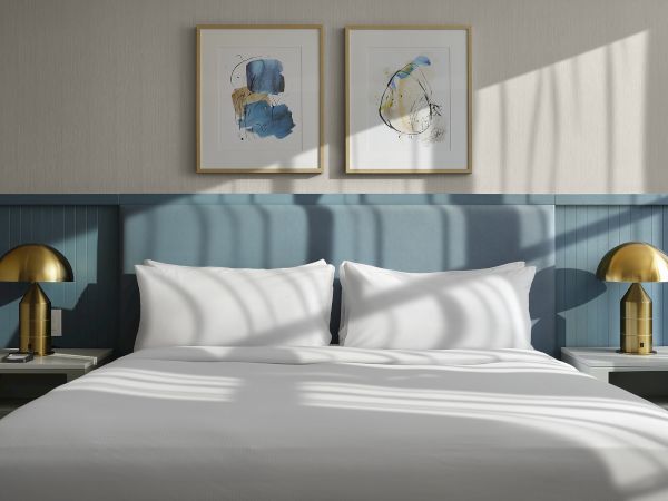 A modern hotel room with a white bed, brass lamps, and two blue abstract paintings on the wall.
