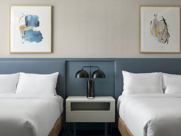 A hotel room with two white beds, a bedside table and twin lamps, and two framed abstract blue paintings on the wall.