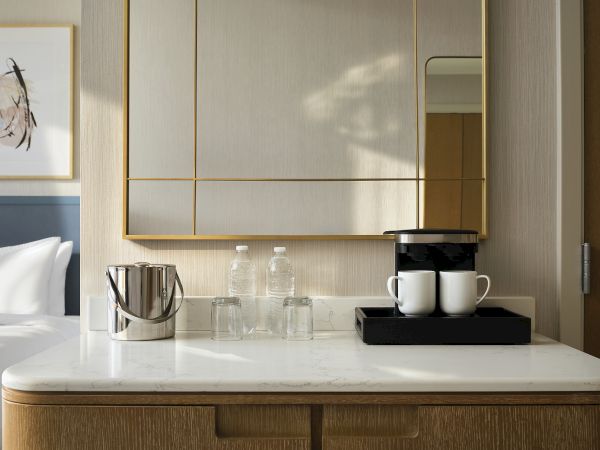 A stylish hotel room coffee station on a marble counter with a kettle, cups, and bottled water beside a framed artwork.
