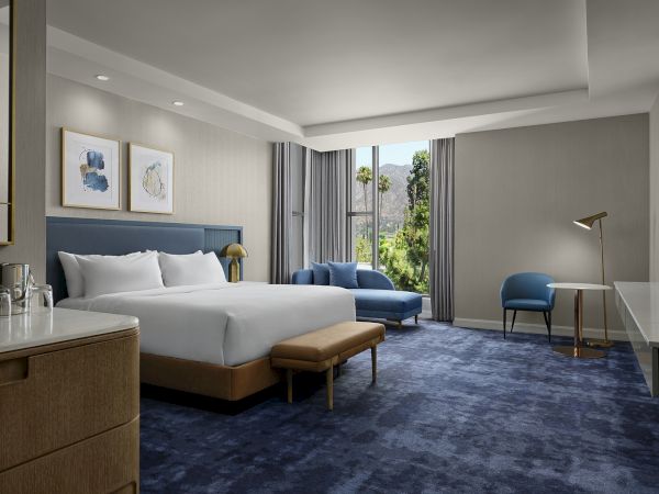 A modern hotel room with a king bed, blue accents, curtains, a desk, and a view of mountains and greenery.
