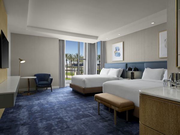 A modern hotel room with two beds, blue accents, large windows, curtains, and a seating area.