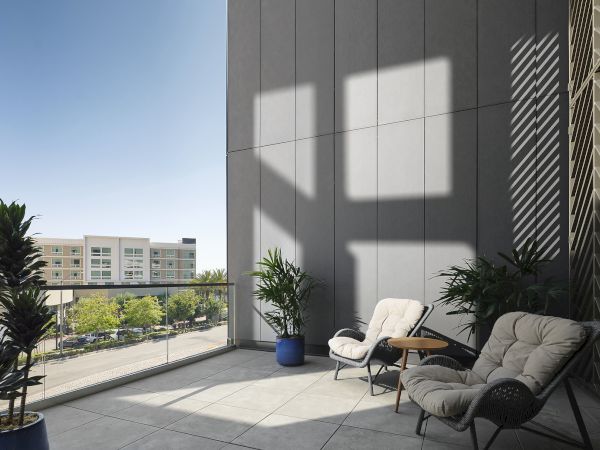 Sunlit apartment balcony with potted plants, lounge chairs, and a small table against a modern gray building wall.