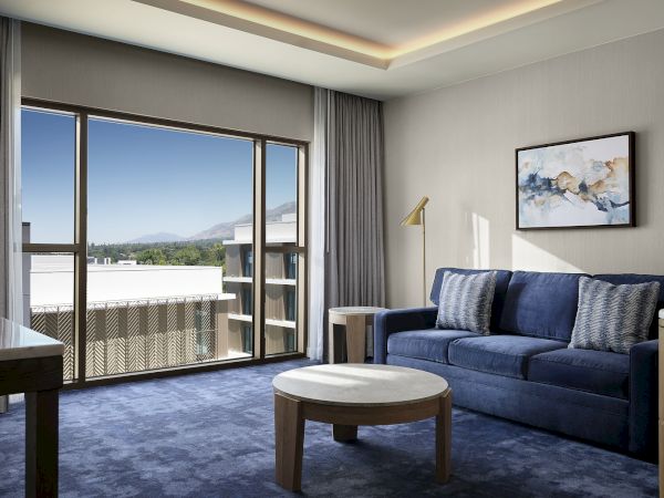 A modern living room with a blue sofa, round coffee table, framed art, and large sliding doors opening to a balcony with mountain views.