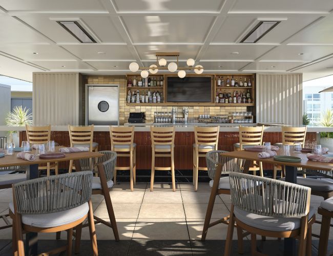 A modern outdoor patio bar with stacked bottles, a glassware display, and neatly set tables with chairs and lounge seating.