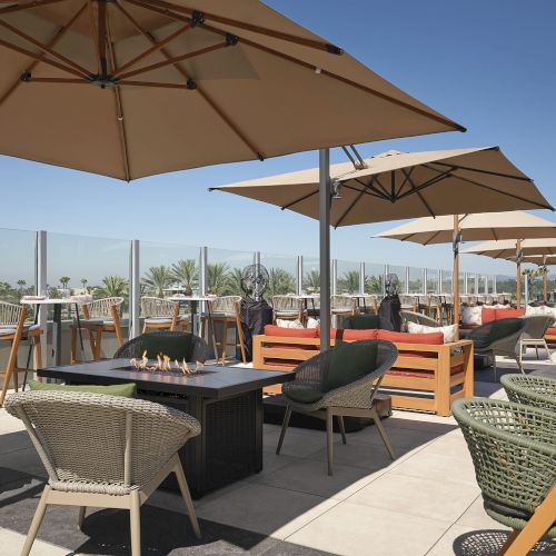 A sunny rooftop patio with wicker chairs, wooden tables, and a gas fire pit under large tan umbrellas.