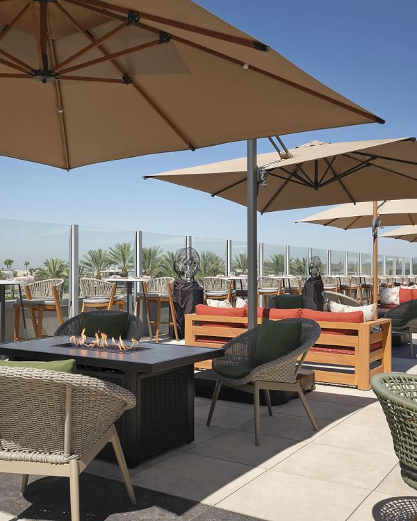 A sunny rooftop patio with wicker chairs, wooden tables, and a gas fire pit under large tan umbrellas.