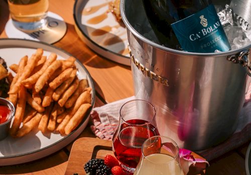 A wine bottle chilling in a silver bucket, assorted cocktails, a plate of fries with dip, and a wooden board topped with berries and sliced fruit.