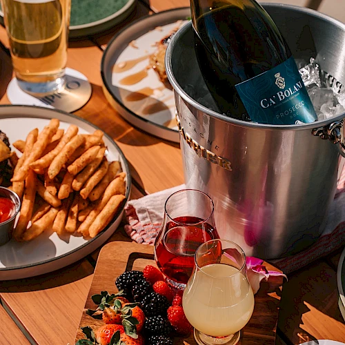 A wine bottle chilling in a silver bucket, assorted cocktails, a plate of fries with dip, and a wooden board topped with berries and sliced fruit.